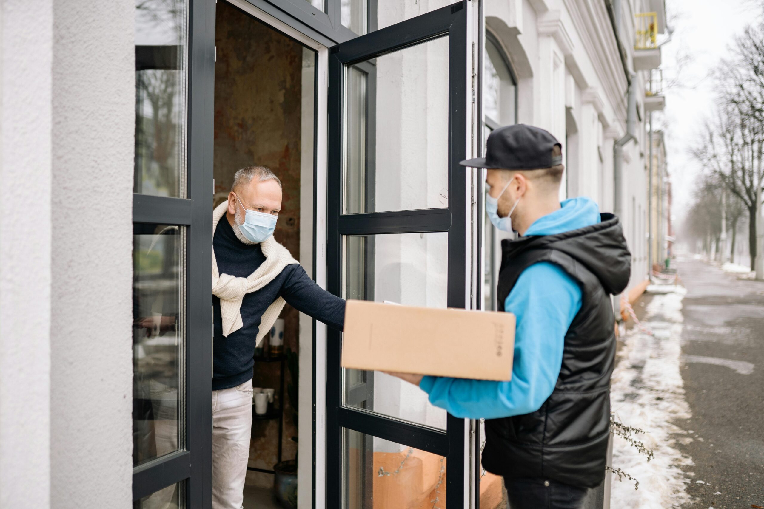 A delivery person hands a parcel to a senior wearing a mask at home, illustrating contactless delivery.